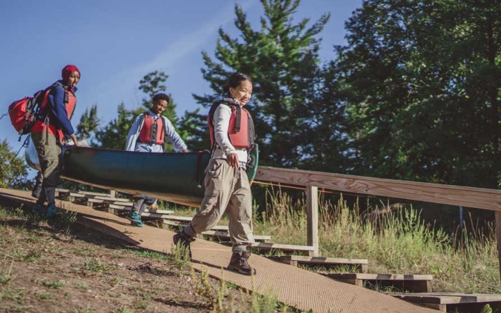 Three people wearing life jackets carry a canoe down a concrete slope, presumably toward water. 